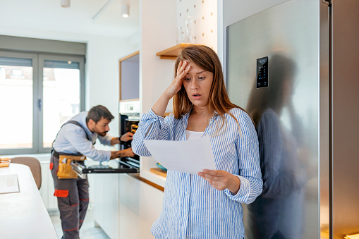 Young Frustrated Woman Looking At Huge Bill From Plumber Standing In Kitchen