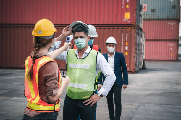 situação de crise epidêmica da doença de coronavirus covid-19, trabalhador de transporte de contêineres com escaneamento corporal de febre por termômetro escaneando no estaleiro. corona-virus covid19 prevenção do novo conceito normal - jarda - fotografias e filmes do acervo