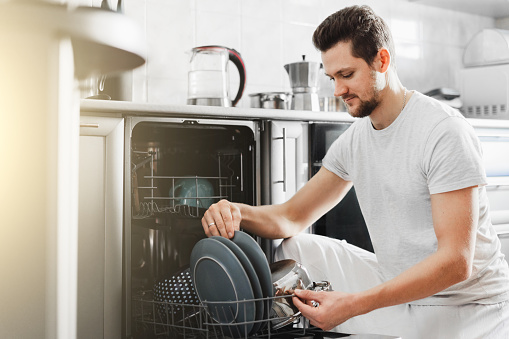 Handsome man unloading dishwasher and enjoying in housework. Husband doing chores in kitchen.