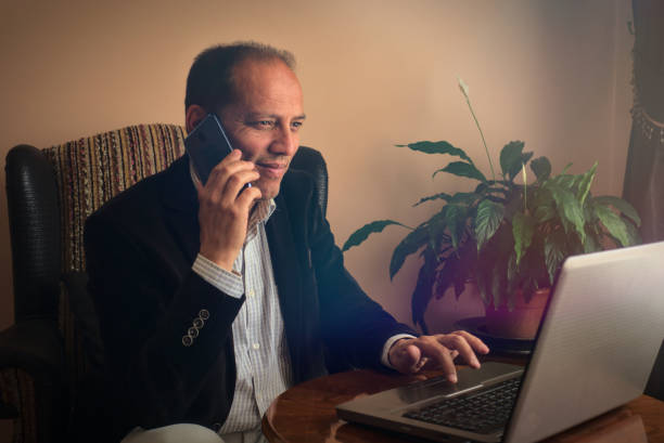 Happy and smiling senior man sitting on armchair phoning to a friend at home stock photo
