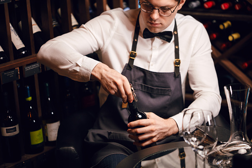 Young handsome sommelier pulling cork out of a bottle using a sommelier knife. Cropped photo of a waiter opening red wine bottle with corscrew in restaurant