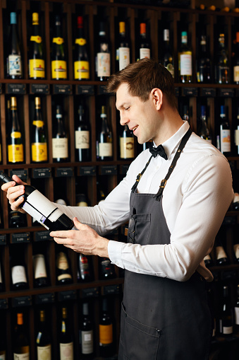Handsome and young elegant brown haired cavist or wine seller with a bottle of wine on the background of dark wine house with shelves of bottles with alcohol.
