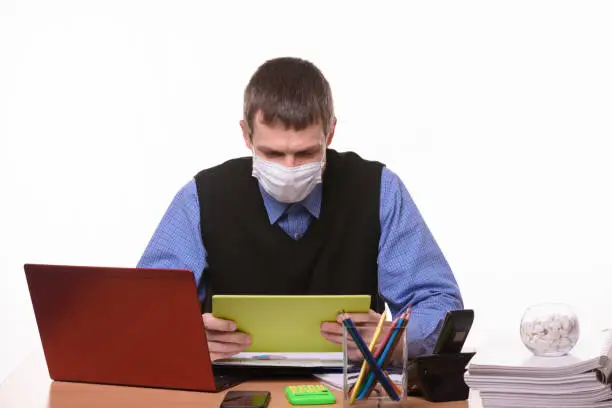 Photo of Office worker in a medical mask at the workplace looks at the tablet screen