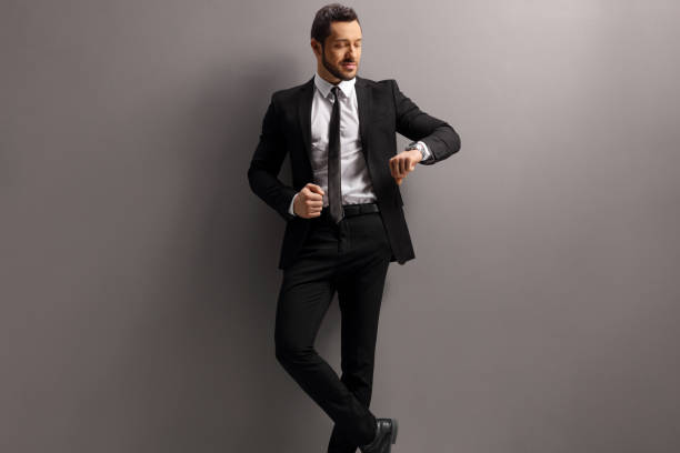 istock Handsome man in a suit checking his watch and leaning on a gray wall 1226061528