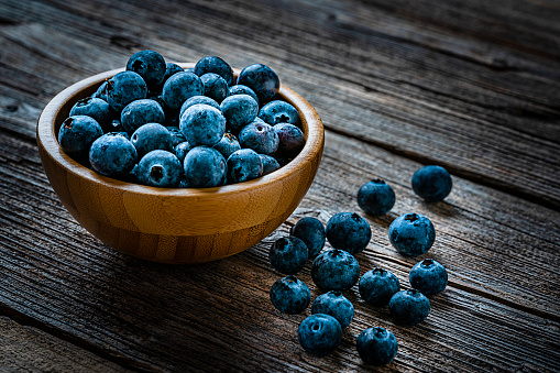 Healthy food: ripe organic blueberries in a bowl shot on rustic wooden table. Some berries are out of the bowl scattered on the table. Predominant colors are blue and brown. High resolution 42Mp studio digital capture taken with Sony A7rII and Sony FE 90mm f2.8 macro G OSS lens