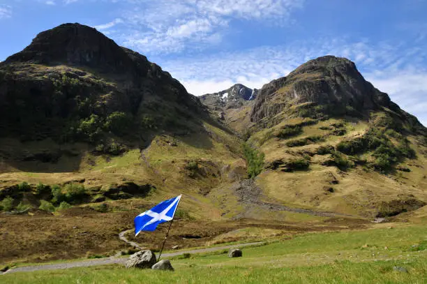 A Scottish flag flattering in the wind with Three sisters mountain range in the background (named Beinn Fhada, Gearr Aonach, and Aonach Dubh). Situated in Glencoe, Scotland's most famous and most scenic Highland glen.