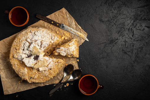 fresh Napoleon cake with a cut piece, knife and spoons on kraft paper with two cups of tea for two on a dark concrete surface. top view. artistic mockup with copy space