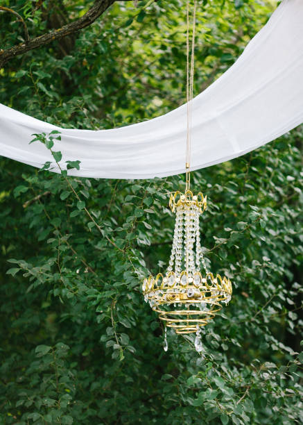 Modern decor, celebration outdoor design and style. Destination elopement festive concept. Wedding arch made of white cotton fabric, decorated with a chandelier against a green forest background.