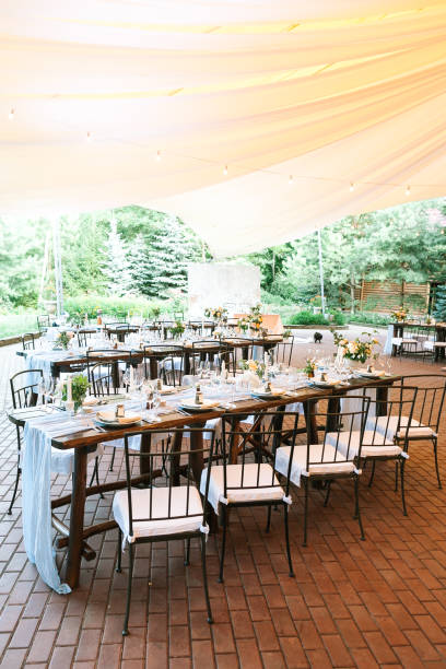 istock Vertical frame. General view of the rows of prepared for reception and beautifully decorated wooden tables with flowers and food sets wedding tables under a large tent 1226073344