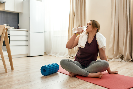 Young curvy woman in sportswear drinking water while sitting after exercising on a yoga mat at home. Determination, will power, sport concept. Horizontal shot