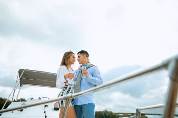 Fashionable stylish standing young couple posing on luxury motorboat yacht on blue sky with clouds and sea landscape background in daylight view from the water stock photo