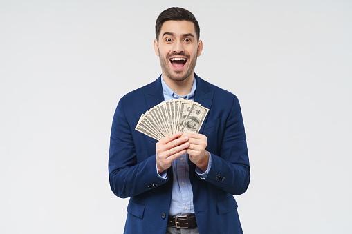 Young man holding fan of dollars in both hands, screaming with joy, winning money in lottery, feeling excited, isolated on gray background