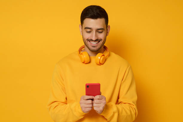 istock Young trendy man wearing sweatshirt and wireless headphones, smiling while looking at content on phone screen, isolated on yellow background 1226086680