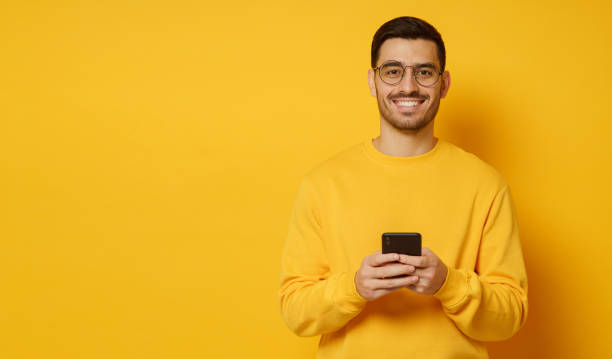 istock Horizontal banner of young man in eyeglasses, smiling happily while looking at camera after communication with friends on phone, isolated on yellow background, copy space on left 1226086689