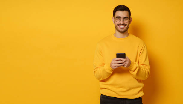 Horizontal banner of man looking happily at camera, holding cellphone in hands, communicating with friends, isolated on yellow background, copy space on left