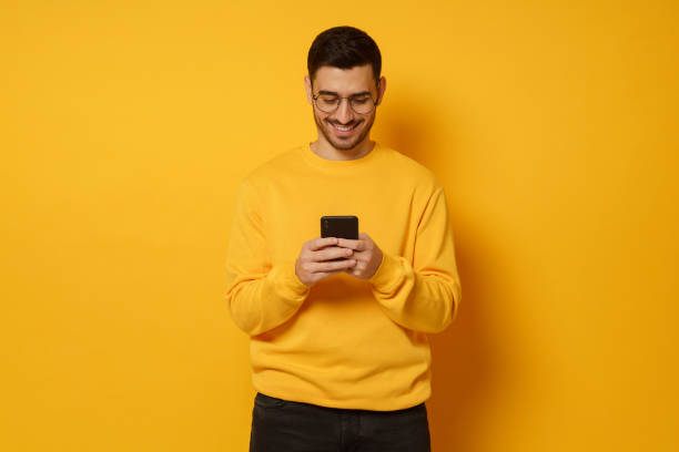 istock Young handsome man exchanging text messages with friends via smart phone, having fun, wearing bright sweatshirt, isolated on yellow background 1226086699