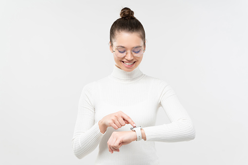 Young woman wearing eyeglasses and wristwatch, checking if she is in time, touching screen, smiling happily, isolated on gray background