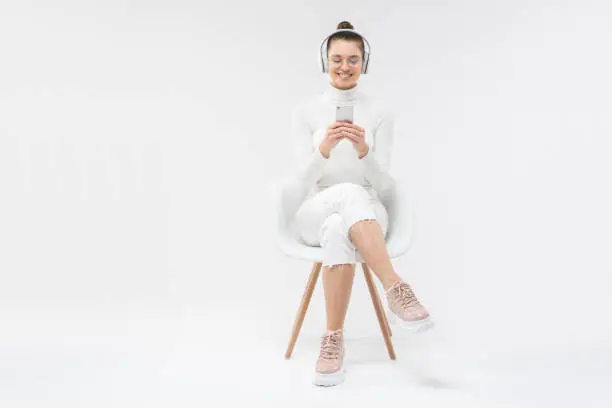 Photo of Young girl sitting on white chair with crossed legs, wearing glasses and wireless headphones, choosing tracks from phone, isolated on gray background