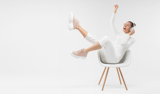 Horizontal banner of young happy girl listening to music in white chair, wearing wireless headhones, isolated on gray background with copy space in studio