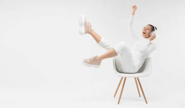 Photo of Horizontal banner of young happy girl listening to music in white chair, wearing wireless headhones, isolated on gray background with copy space in studio