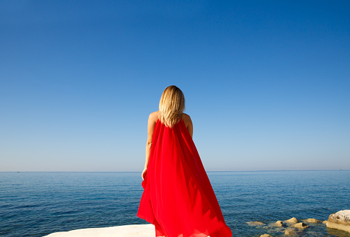 Young red-haired girl in red ball gown dances incendiary dance on green meadow against blue evening summer cloudy sky.