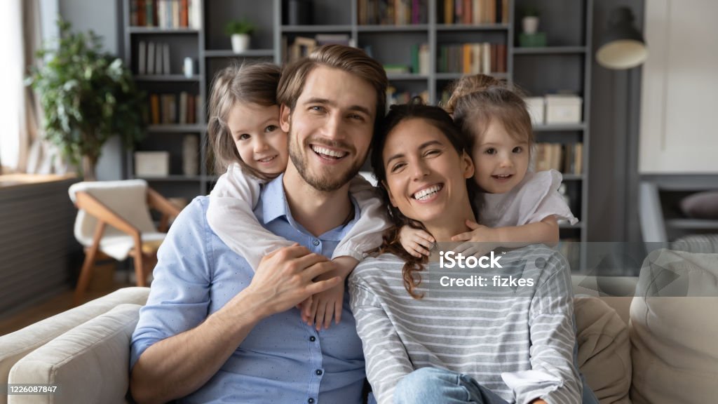 Portrait of young family with small daughters at home Portrait of young Caucasian family with small daughters pose relax on sofa in living room, smiling little girls kids hug embrace parents, show love and gratitude, rest on couch at home together Family Stock Photo