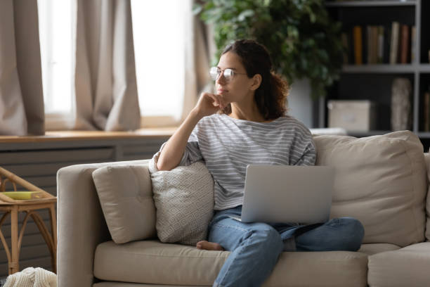 Young woman distracted from computer looking in distance Dreamy young Caucasian girl in glasses sit on couch in living room hold laptop look in window distance dreaming, thoughtful female distracted from computer work dreaming or thinking at home Distracted stock pictures, royalty-free photos & images