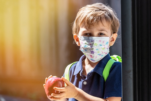 Caucasian Little schoolboy holding an apple wearing a protective mask and a backpack