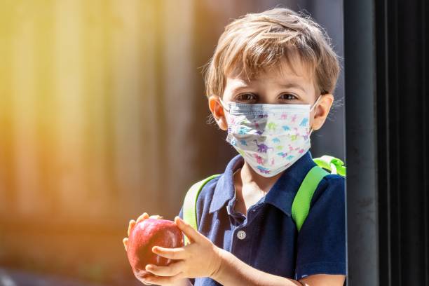 little schoolboy holding an apple wearing a protective mask and a backpack - little boys preschooler back to school backpack imagens e fotografias de stock