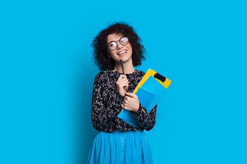 Charming curly haired student holding some books while posing in a dress on blue wall with blank space