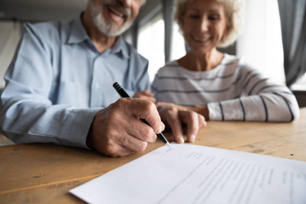 Close up focus on wrinkled male hand signing paper document. Close up focus on wrinkled male hand signing paper document. Smiling elderly mature family couple putting signature on leasing or medical insurance contract, purchase agreement with real estate agent legalization photos stock pictures, royalty-free photos & images