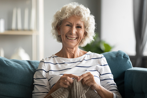 Happy elderly mature beautiful hoary grandmother relaxing on comfortable sofa, knitting warm sweater with woolen threads. Smiling middle aged woman enjoying free hobby weekend time alone at home.