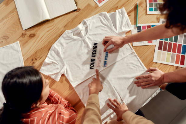 scrivere che vende. persone creative che provano gli adesivi con testo, mentre discutono del logo e del design della t-shirt. giovane uomo e donna che lavorano insieme alla t-shirt personalizzata, azienda di stampa di abbigliamento - textile print foto e immagini stock