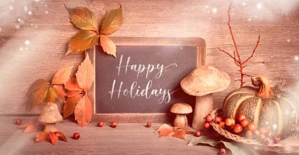 Text "Happy Holidays" on blackboard. Autumn traditional decorations, toned panoramic image. Fall leaves, berries, mushrooms, pumpkins, flowers on wood. Thanksgiving still life.