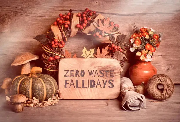 Text "Zero waste Fallidays" on wooden board. Autumn traditional decorations, toned image. Fall wreath with berries, mushrooms, pumpkins, flowers on wood.