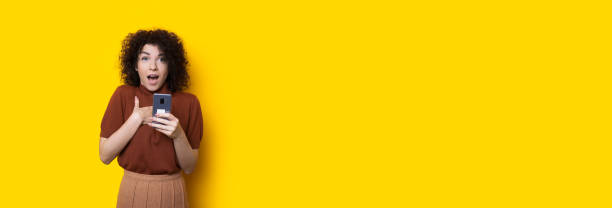 istock Curly haired lady pointing to herself while holding a mobile and posing on a yellow wall with blank space 1226096076