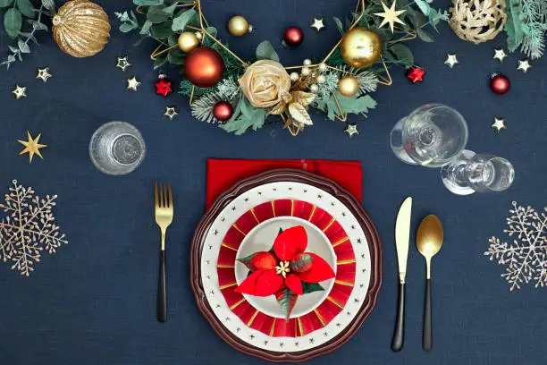 Christmas table setting in gold, burgundy and classic blue colors. Flat lay, top view on decorative table layout, golden cutlery, white plates with stars. Traditional Xmas decor on classic blue linen