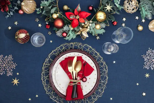 Christmas table setting in gold, burgundy and dark blue colors. Flat lay, top view on decorative table layout with golden cutlery, white plates with stars, traditional decorations on dark blue linen