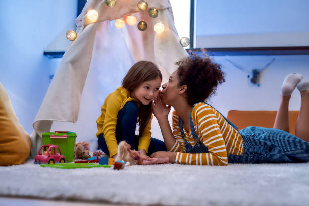 afrikanische amerikanische frau baby sitter unterhaltsam kaukasischen niedlichen kleinen mädchen. sie klatschen und erzählen geheimnisse, die im kinderzimmer sitzen - babysitter stock-fotos und bilder