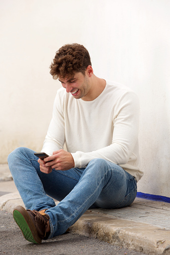 Full body side portrait of laughing man sitting on ground with mobile phone