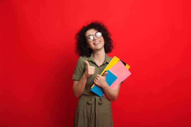 istock Curly haired caucasian student is holding some books and gesturing the like sign while posing on a red background 1226108877
