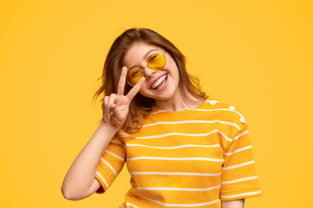 Cheerful young hipster gesturing V sign Excited young woman in bright outfit and sunglasses smiling and keeping two fingers near eye against yellow background peace sign gesture photos stock pictures, royalty-free photos & images