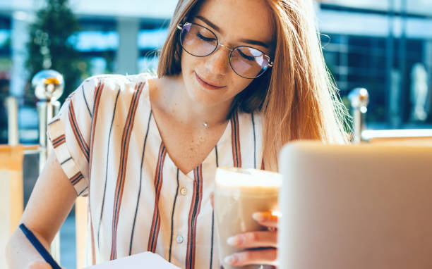 istock Ginger business lady with eyeglasses is writing something while using a laptop and drinking a cocktail at the coffee shop 1226121289