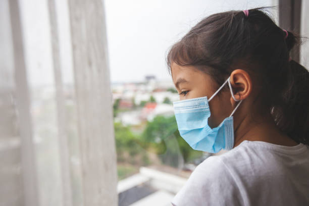 fille asiatique portant le masque de protection regardant dehors par la fenêtre et rester à la quarantaine à la maison du coronavirus covid-19 et la pollution de l’air pm2.5. - mirror reflection blues glass photos et images de collection