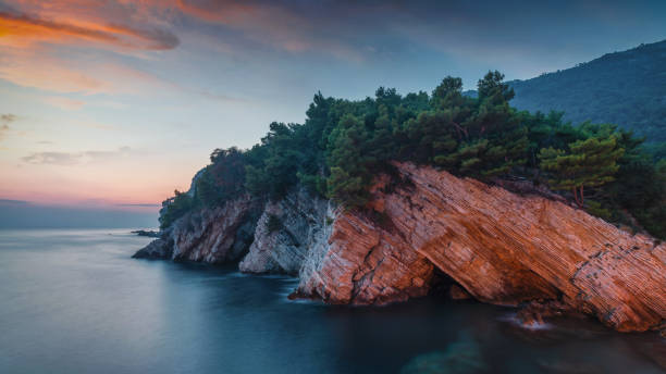Panorama of the beautiful rocky adriatic sea coast of Petrovac under moody colorful sunset twilight. Illumimnated Cliffs at Petrovac, located on the coast between the cities Budva and Bar. Petrovac, Montenegro, Southeast Europe, Europe.