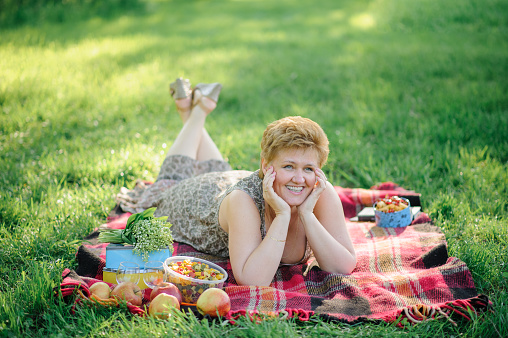 The senior woman lies belly on a plaid and poses for the camera in the park on the nature.