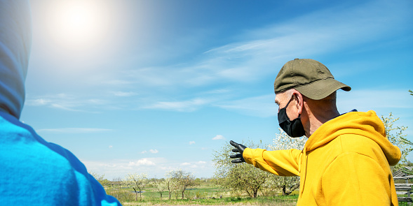 farmer in black mask and inverted baseball cap points to field standing by person at vegetable garden on spring day closeup