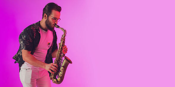 Young caucasian jazz musician playing the saxophone on gradient pink studio background in neon light. Concept of music, hobby, festival. Joyful, cheerful attractive guy. Colorful portrait of artist.