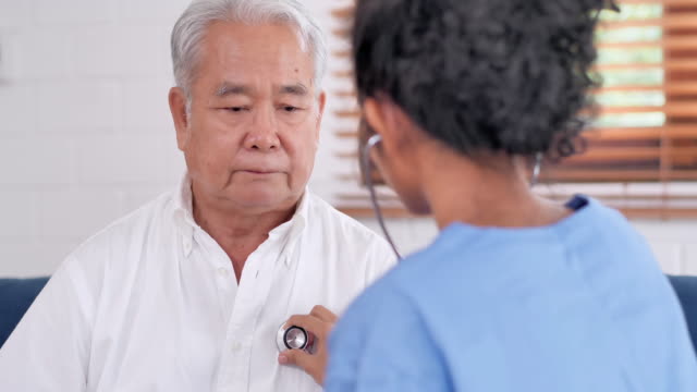 Young black female nurse doctor checking heart to asian older men to prevent epidemics of Coronavirus or Covid-19 at home.Medical,Caretaking,Care,Retirement,Volunteer,Charity,Education,Nursing home,Caregivers at Home,Medicine and health care