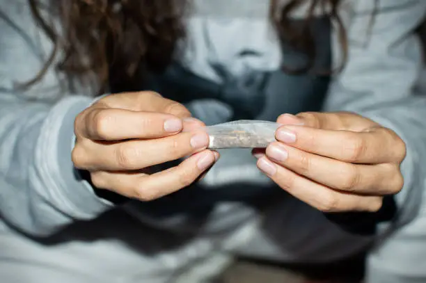 Photo of Young woman in tracksuit rolling a marijuana joint in the street at night. Details of hands rolling cannabis cigarette.